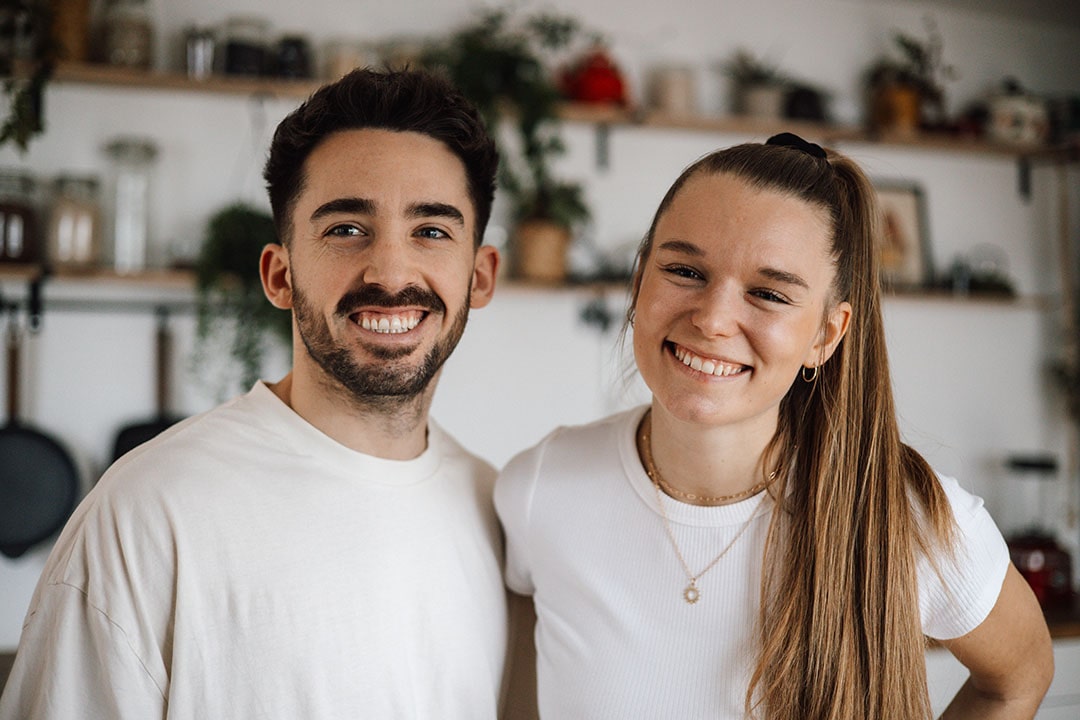 Two people smiling in a kitchen with shelves and plants.